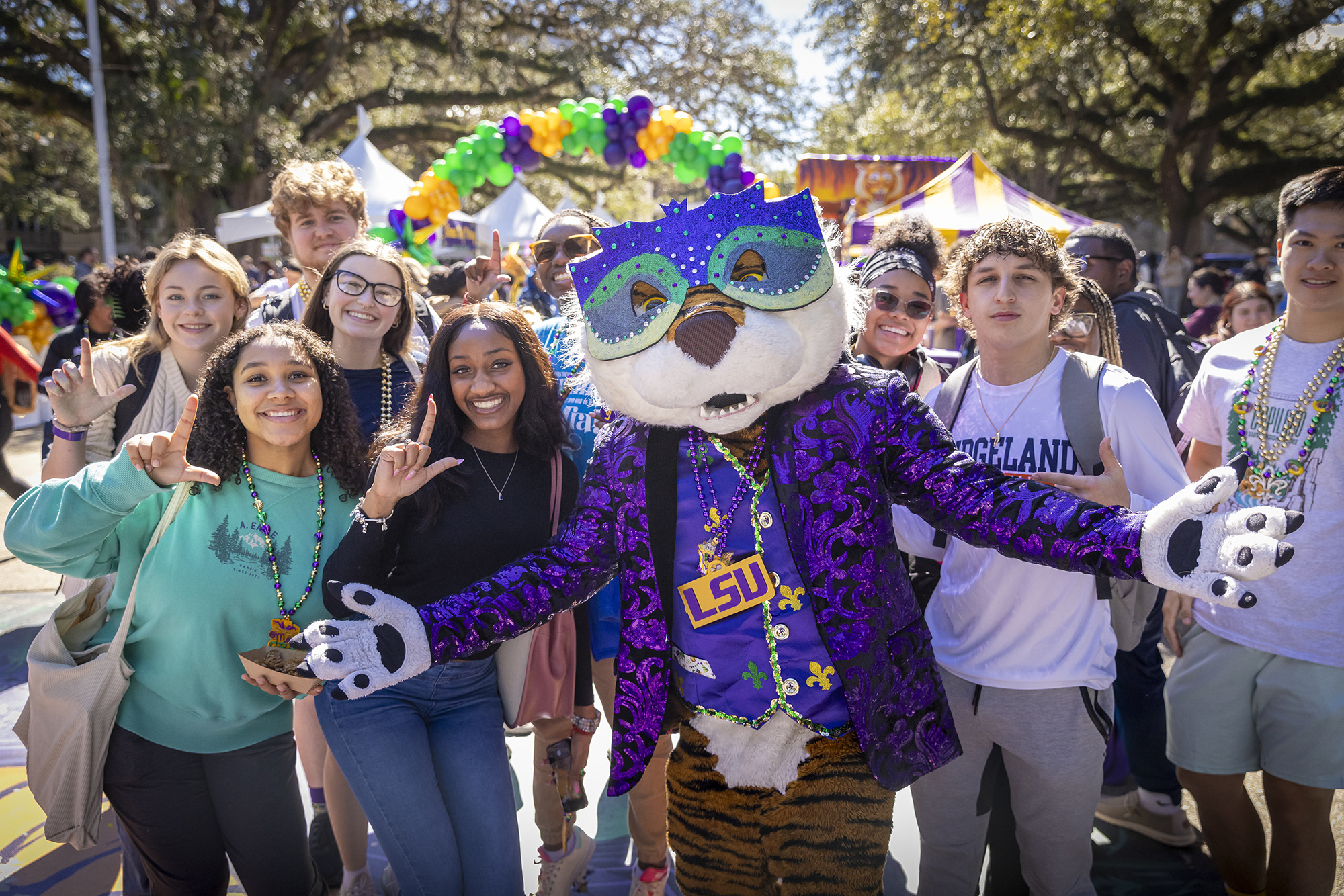 Mike mascot dressed in colorful attire and a Mardi Gras mask poses with students