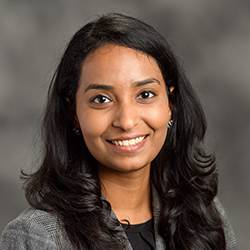 A close-up of a person with long dark hair smiling and wearing a suit (Ripinka Patil headshot)