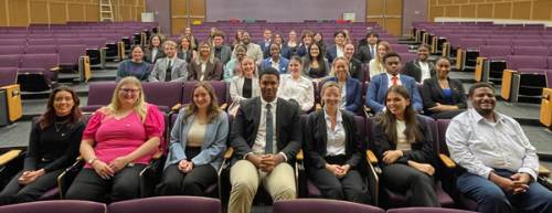 Delta Sigma Pi members sit in chairs in the BEC Auditorium and look straight ahead and smile at the camera.