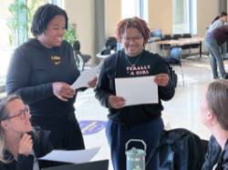 Two students stand above a table where professionals are seated. They have a lively conversation.