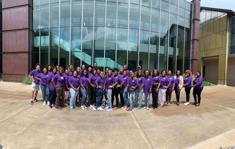 a group of bootcamp participants stand outside the Business Education Complex at LSU, they are wearing purple polos and jeans