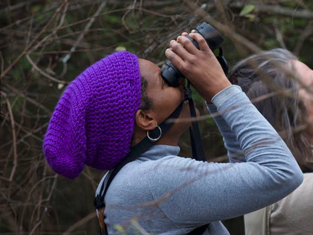 a woman wearing a purple beanie looks up at the sky through binoculars