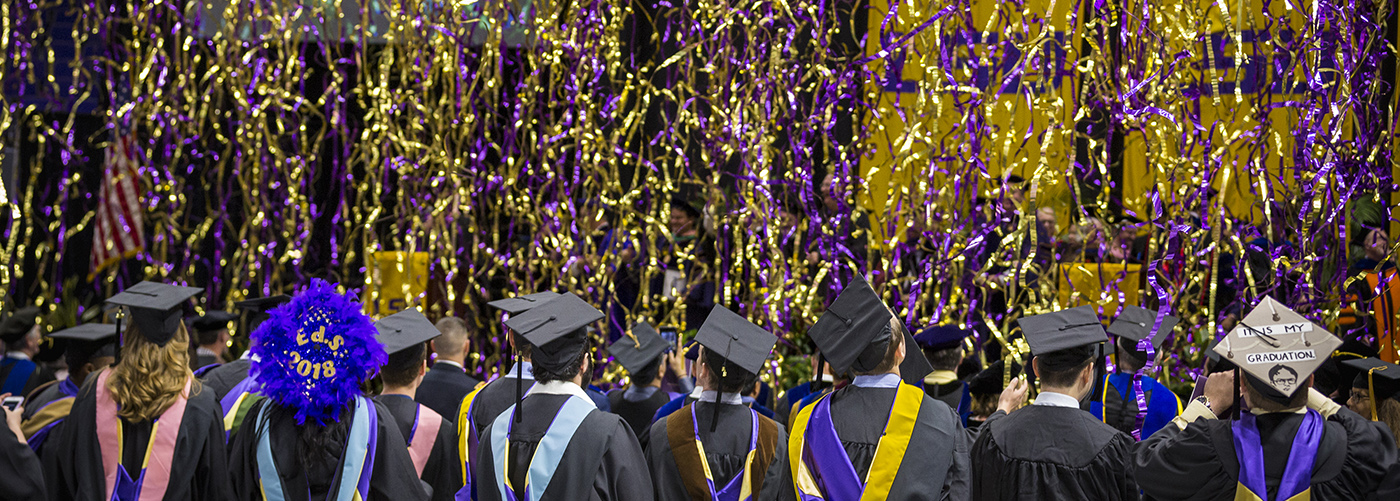 Spring graduates in regalia with purple and gold streamers falling from the ceiling.