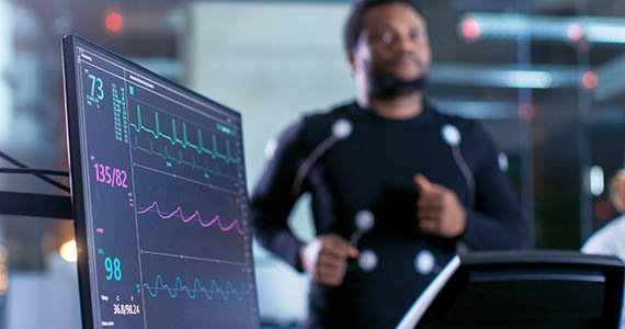 Photo of a man running on a treadmill with leads on his chest with a monitor showing his heartrate.