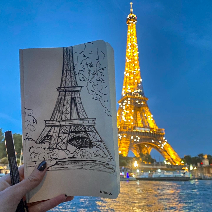 student holds up drawing of Eiffel Tower next to actual Eiffel Tower
