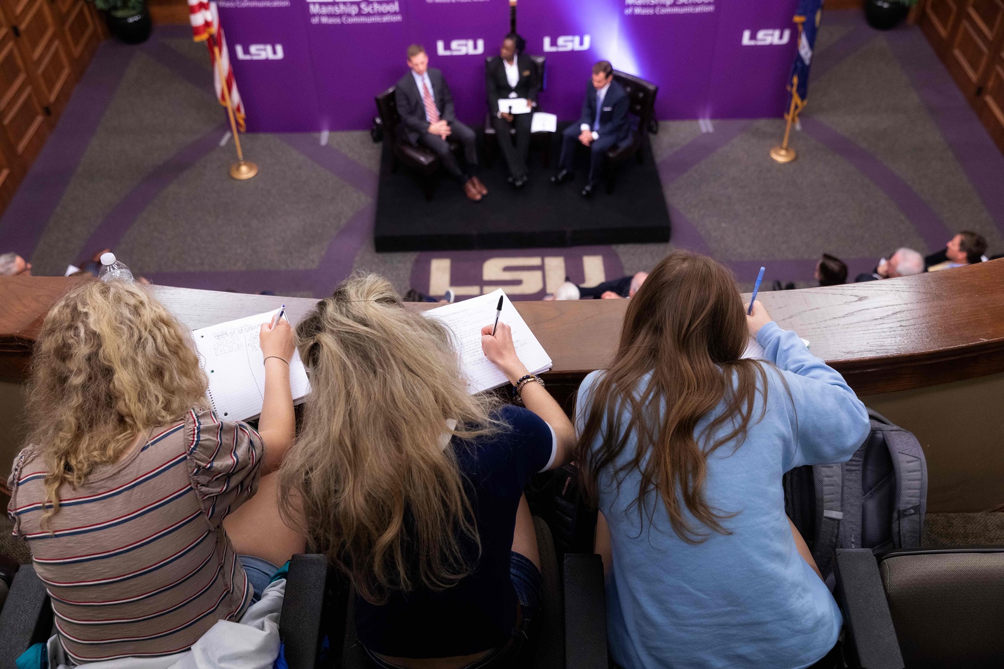 three female students write on pieces of paper on the top balcony while overlooking the speakers in the forum