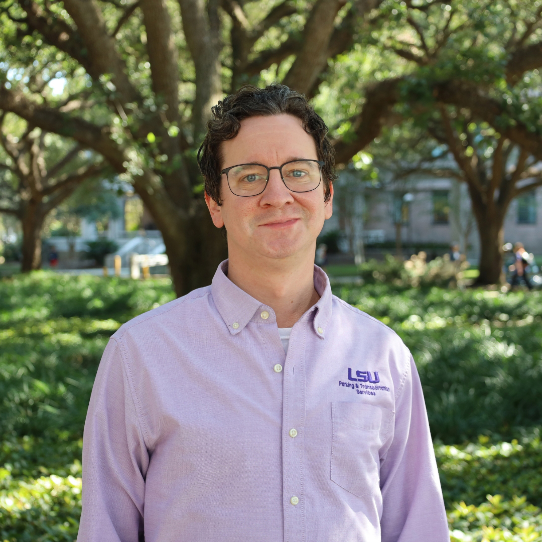 Man in purple shirt smiling and standing in front of a tree and bushes
