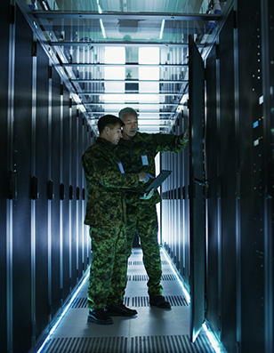 Two military men is camouflage holding a laptop in a hallway filled with computer servers.